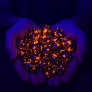 May include: A cluster of glowing, speckled stones held in cupped hands. The stones have a dark base colour with bright orange and yellow spots, illuminated against a dark blue background. The stones appear to be egg-shaped.