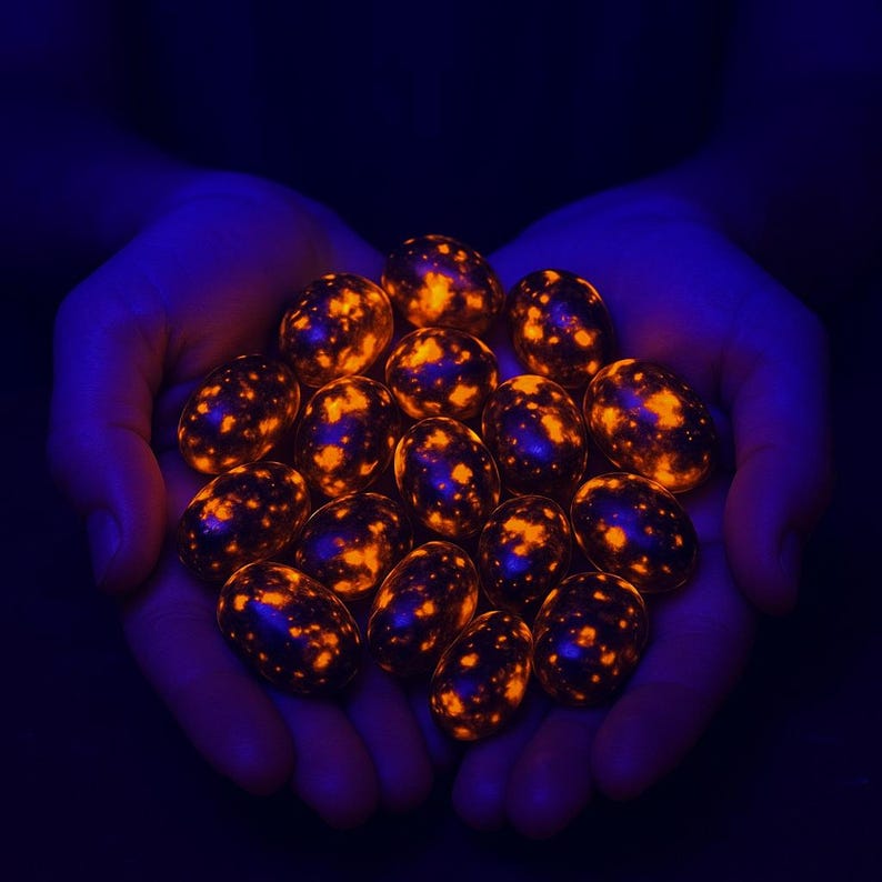 May include: A cluster of glowing, speckled stones held in cupped hands. The stones have a dark base colour with bright orange and yellow spots, illuminated against a dark blue background. The stones appear to be egg-shaped.