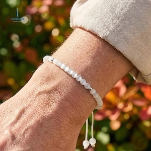 May include: A white beaded bracelet with a silver accent is worn on a wrist. The bracelet is made of small, round, white beads strung together. The bracelet is adjustable with a white string. The background is blurred with green and brown tones.
