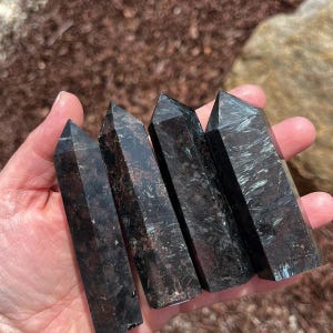 May include: Five black and green gemstone points, also known as black tourmaline, are arranged in a row in a hand. The points are smooth and polished.