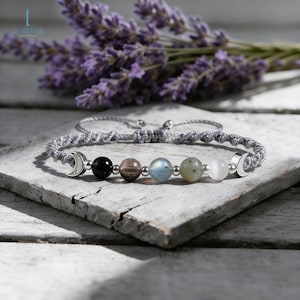 May include: A gray macrame bracelet with a series of colorful gemstone beads and silver crescent moon charms. The bracelet features black, brown, blue, and white beads. The background includes lavender and a wooden surface.