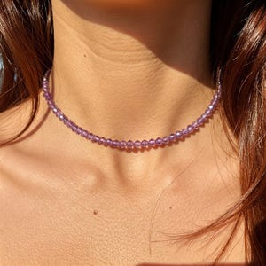 May include: A delicate choker necklace composed of small, faceted lavender beads. The necklace sits close to the neck, creating a subtle and elegant look. The beads catch the light, adding a touch of sparkle to the design.