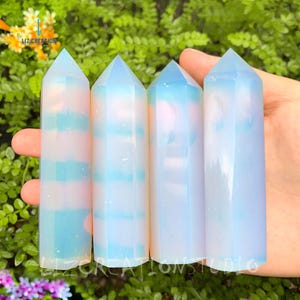 May include: Four iridescent blue and white opal crystal points, also known as angel aura quartz, are arranged in a row. The crystals are smooth and polished, with a translucent appearance. The crystals are held in a hand, with the background blurred.