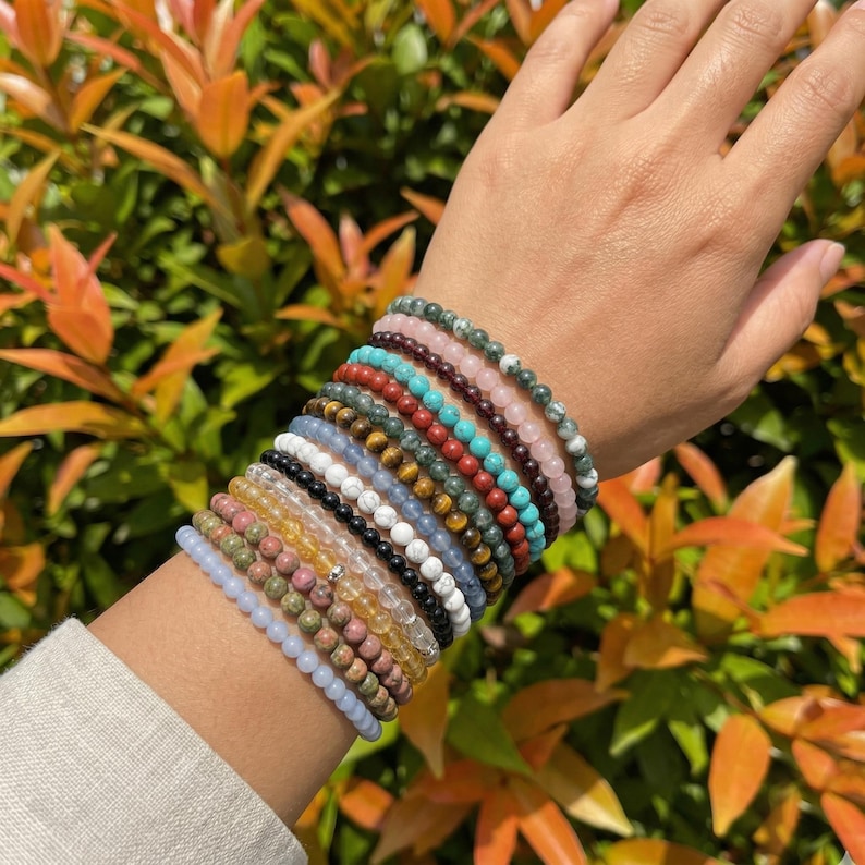 May include: A collection of beaded bracelets in various colors, including pink, blue, green, and brown. The bracelets are stacked on a wrist, showcasing the variety of gemstones and textures. The background features vibrant green and orange foliage.