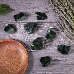 Puede incluir: Una colecci&oacute;n de rocas de obsidiana verde oscuro con bordes irregulares, dispersas sobre una superficie de madera. Tambi&eacute;n hay un cuenco de madera y ramitas de lavanda secas. Las rocas var&iacute;an en tama&ntilde;o y forma.