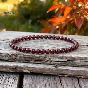 May include: A beaded bracelet made of small, round, deep red gemstones. The bracelet is resting on a weathered wooden surface, with a blurred background of green and orange foliage. The text "LIZ CREATION" is visible in the upper left corner.