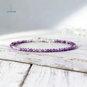 May include: A delicate bracelet featuring small, faceted purple beads. The beads are strung together with a silver clasp. The bracelet is displayed on a white wooden surface. The brand name "LIZ CREATION" is visible in the upper left corner.