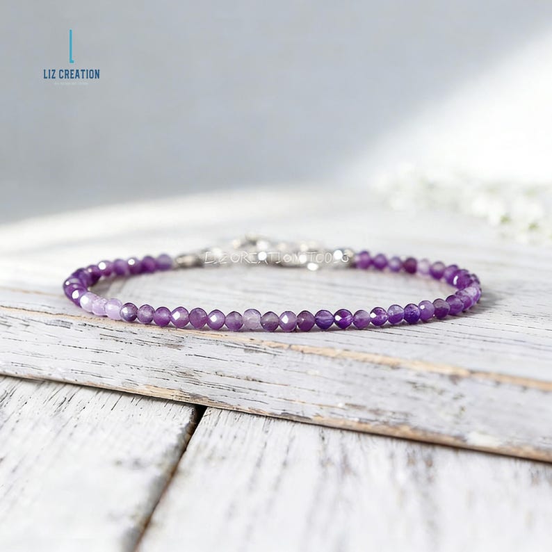 May include: A delicate bracelet featuring small, faceted purple beads. The beads are strung together with a silver clasp. The bracelet is displayed on a white wooden surface. The brand name "LIZ CREATION" is visible in the upper left corner.