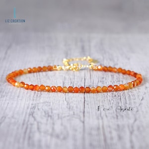 May include: A delicate necklace made with small, orange fire agate beads. The necklace is strung on a gold chain and has a simple, elegant design.  "Fire Agate" is written in white text below the necklace.
