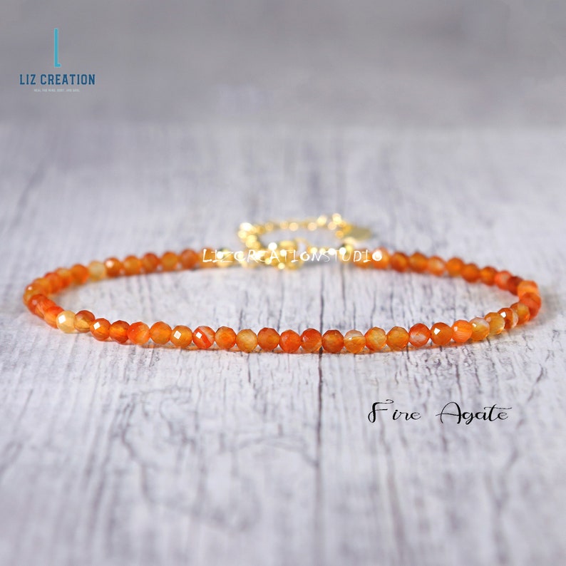 May include: A delicate necklace made with small, orange fire agate beads. The necklace is strung on a gold chain and has a simple, elegant design.  "Fire Agate" is written in white text below the necklace.