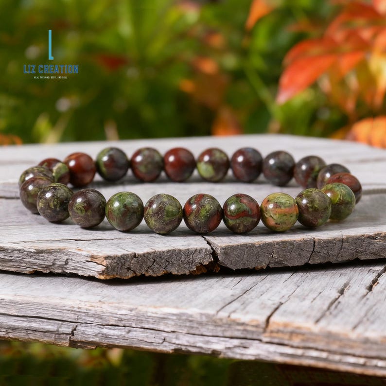Puede incluir: Una pulsera de cuentas redondas y pulidas en tonos verdes, marrones y rojos. La pulsera se muestra sobre una superficie de madera envejecida. El texto "Liz Creation" es visible en la esquina superior izquierda.