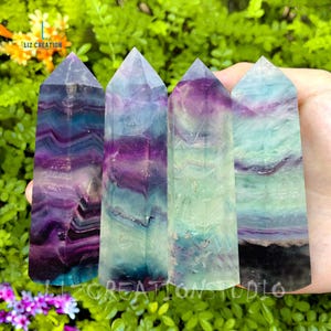 May include: Four fluorite crystal points with green, purple and blue banding. The crystals are arranged in a row and are being held by a hand. Liz Creation Studio is printed on the bottom of the image.