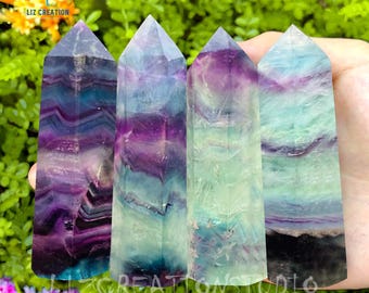 May include: Four fluorite crystal points with green, purple, and blue banding. The crystals are arranged in a row and are being held by a hand. Liz Creation Studio is printed on the bottom of the image.