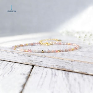 Puede incluir: Delicada pulsera de cuentas con pequeñas piedras preciosas redondas rosas y grises intercaladas con cuentas doradas. La pulsera se exhibe sobre una superficie de madera blanca. El cierre y la cadena son dorados. El nombre de la marca "LIZ CREATION" está en la esquina superior izquierda.