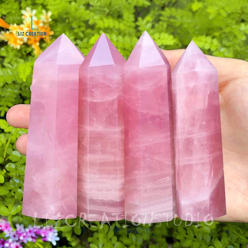 K&ouml;nnte beinhalten: Vier polierte Rosenquarz-Obelisken. Jeder Obelisk hat eine hellrosa Farbe mit einer spitzen Spitze. Die Obelisken werden in einer Hand vor einem Hintergrund aus gr&uuml;nem Laub gehalten. Der Text "LIZ CREATION" und "LIZ CREATIONS STUDIO" sind sichtbar.