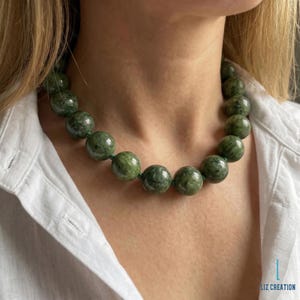 May include: A necklace featuring a single strand of large, round, polished green beads with mottled dark and light green patterns. The beads are uniform in size and strung together, resting against a white collared shirt. The text LIZ CREATION is visible.