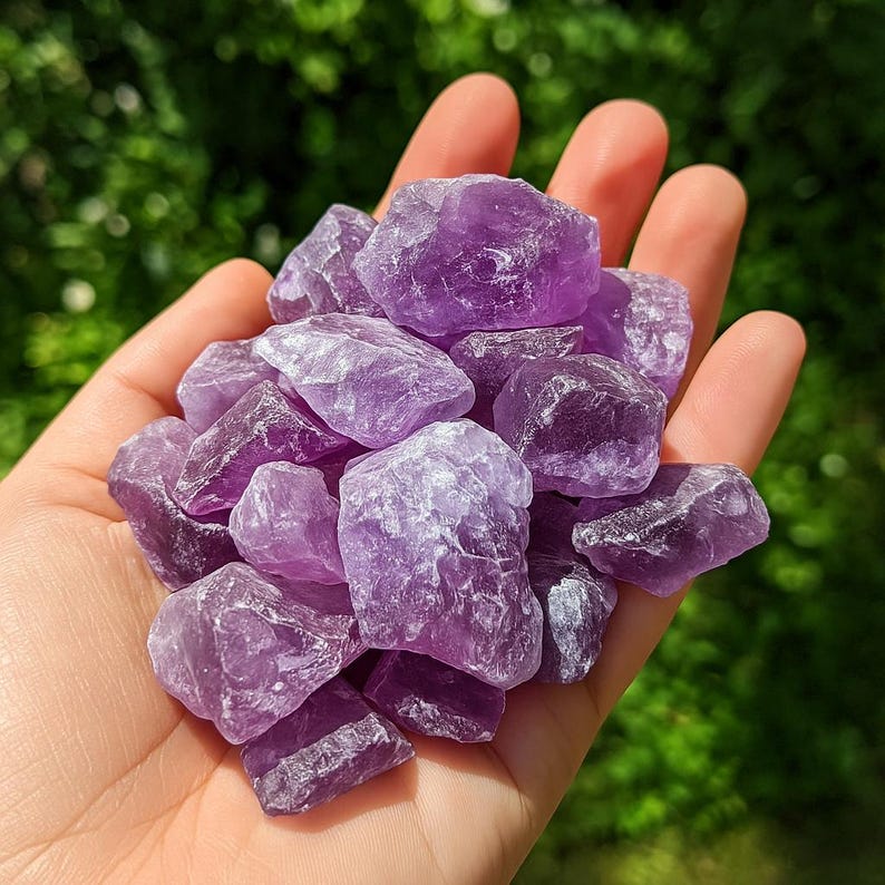 May include: A handful of raw amethyst crystals in varying shades of purple. The rough-cut stones have a natural, textured appearance, with some areas appearing darker than others. The background is a blurred green.
