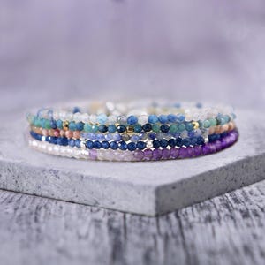 May include: A stack of beaded bracelets in various colors, including blue, purple, white, and green. The bracelets are made of small, faceted beads and are stacked on a gray surface. The bracelets have gold accents.