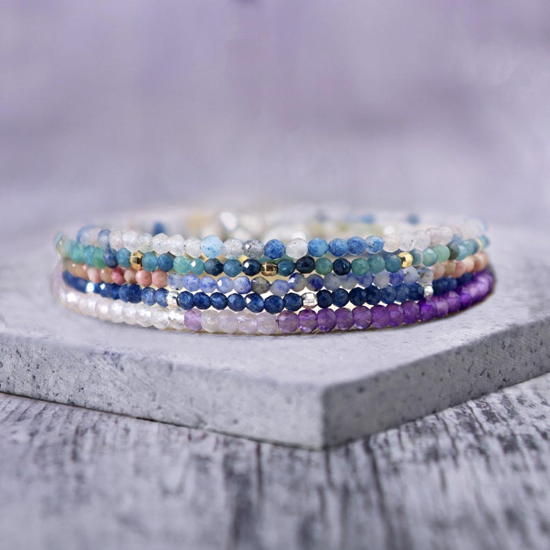 May include: A stack of beaded bracelets in various colors, including blue, purple, white, and green. The bracelets are made of small, faceted beads and are stacked on a gray surface. The bracelets have gold accents.