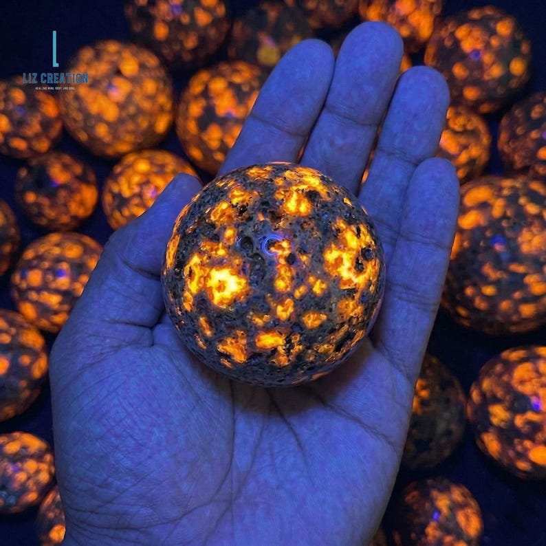 Puede incluir: Una mano sostiene una piedra esf&eacute;rica brillante con una superficie oscura y texturizada e inclusiones de color naranja y amarillo brillante. Hay otras esferas similares en el fondo. La imagen tiene un tinte azul.