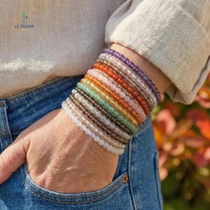 May include: A wrist is adorned with a stack of beaded bracelets in various colors, including purple, orange, green, and white. The bracelets are made of small, round beads. The person is wearing a light-colored shirt and blue jeans. The brand name "LIZ CREATION" is visible.