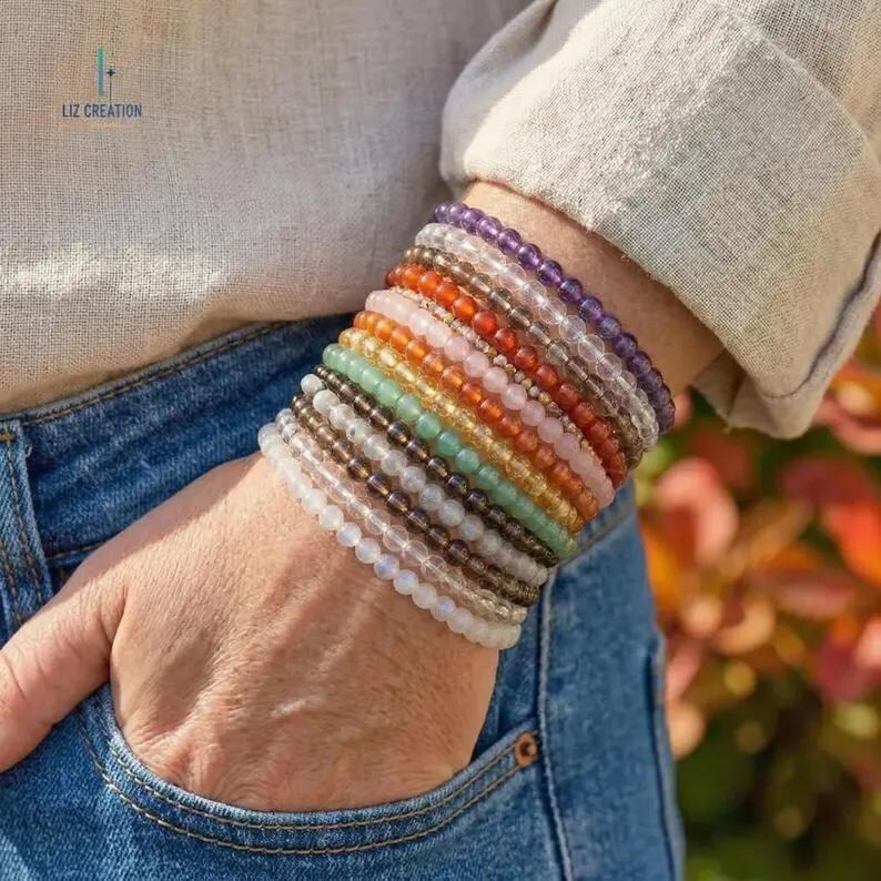 May include: A wrist is adorned with a stack of beaded bracelets in various colors, including purple, orange, green, and white. The bracelets are made of small, round beads. The person is wearing a light-colored shirt and blue jeans. The brand name "LIZ CREATION" is visible.