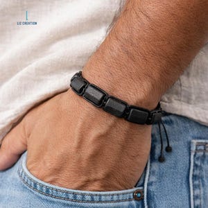 May include: Black beaded bracelet with square-shaped beads, worn on a wrist. The bracelet is made of dark beads strung on a black cord. The person is wearing a light-colored shirt and blue jeans.