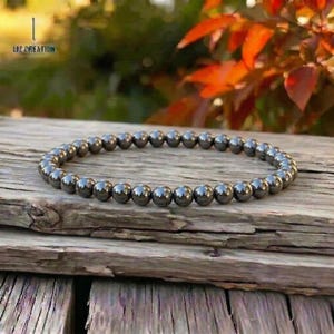May include: A beaded bracelet composed of numerous, small, dark gray, spherical beads. The bracelet is resting on a weathered, wooden surface. The background features blurred greenery and red-orange foliage. The bracelet is a piece of jewelry.