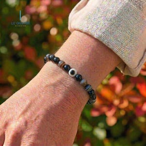 May include: A bracelet made of black, brown, and white beads. The bracelet has a central bead with a white eye design. The bracelet is worn on a wrist. The brand name "LIZ CREATION" is visible in the background.