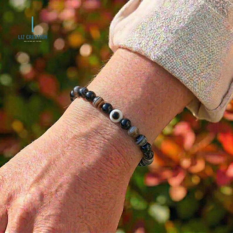 May include: A bracelet made of black, brown, and white beads. The bracelet has a central bead with a white eye design. The bracelet is worn on a wrist. The brand name "LIZ CREATION" is visible in the background.