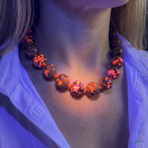 May include: A necklace with large, spherical beads in a speckled orange and dark brown pattern. The beads are strung on an orange cord. The necklace is worn against a white shirt.