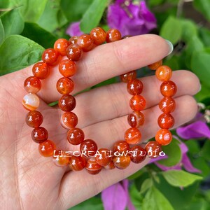 Puede incluir: Dos pulseras hechas de cuentas redondas y pulidas de color naranja rojizo. Las cuentas tienen un aspecto veteado con rayas blancas y naranjas. Las pulseras se sostienen en una mano, con el texto "LIZCREATIONSTUDIO" visible.
