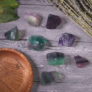 Puede incluir: Una colecci&oacute;n de cristales de fluorita en bruto en tonos verdes, morados y blancos se exhibe sobre una superficie de madera. Tambi&eacute;n hay un cuenco de madera y ramitas de lavanda. Los cristales var&iacute;an en tama&ntilde;o y forma, mostrando texturas naturales.