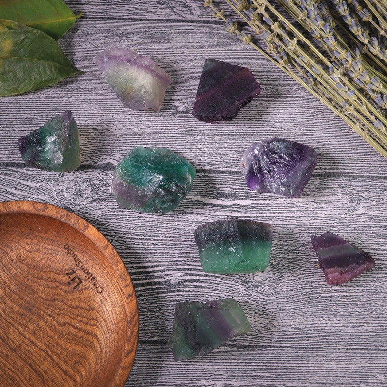 Puede incluir: Una colecci&oacute;n de cristales de fluorita en bruto en tonos verdes, morados y blancos se exhibe sobre una superficie de madera. Tambi&eacute;n hay un cuenco de madera y ramitas de lavanda. Los cristales var&iacute;an en tama&ntilde;o y forma, mostrando texturas naturales.