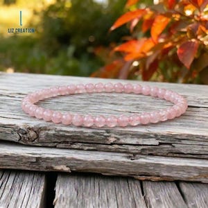 May include: A delicate bracelet composed of small, round, light pink beads. The beads are strung together to form a flexible band. The bracelet is displayed on a weathered, gray wooden surface, with a blurred background of green and orange foliage.
