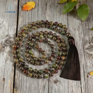 May include: A beaded necklace with a dark brown tassel. The beads are a mix of green, brown, and red hues. The necklace is displayed on a weathered wooden surface, with a few leaves scattered around.