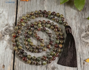 May include: A beaded necklace with a dark brown tassel. The beads are a mix of green, brown, and red hues. The necklace is displayed on a weathered wooden surface, with a few leaves scattered around.