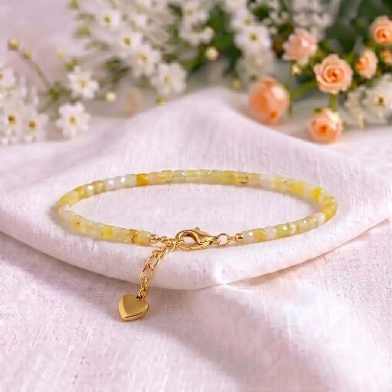 May include: A delicate bracelet featuring small, faceted yellow and white beads strung together. The piece includes a gold-toned metal lobster claw clasp, an adjustable extender chain, and a small, smooth gold-toned heart-shaped charm.