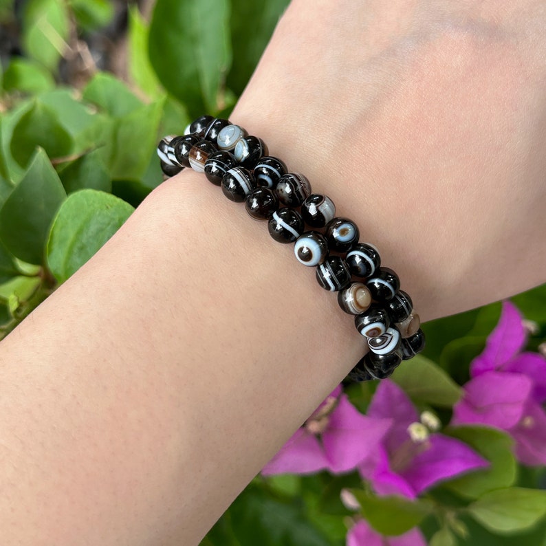 May include: Three black and white beaded bracelets are stacked on a wrist. The beads are round with a pattern of white stripes and eye-like designs. The background features green leaves and purple flowers.