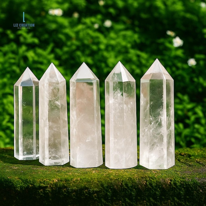 May include: Five clear quartz crystal towers with pointed tops are arranged on a mossy surface against a blurred green background. The crystals are transparent and reflect light, showcasing their internal structures. The Liz Creation logo is in the top left corner.