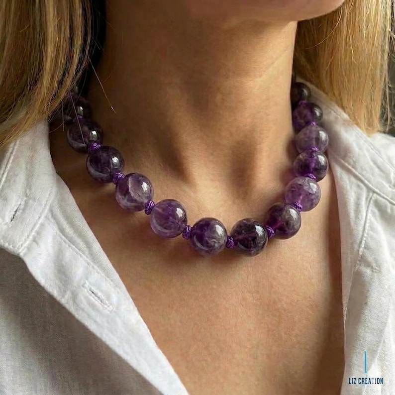 May include: A necklace of large, round amethyst beads in shades of purple, strung on a purple cord. The necklace is worn against a white shirt. The amethyst beads are a striking feature of this jewellery.