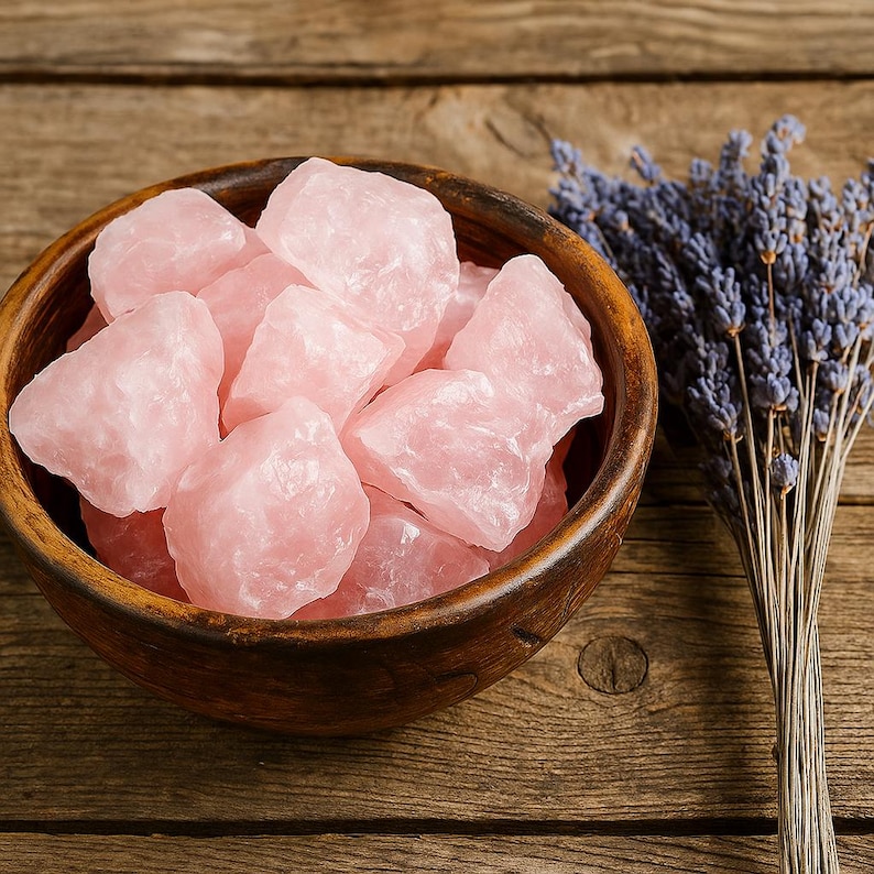 May include: A wooden bowl filled with raw, light pink rose quartz crystals. A bundle of dried lavender with purple flowers sits to the right. The crystals have a rough, natural texture. The bowl is on a wooden surface.