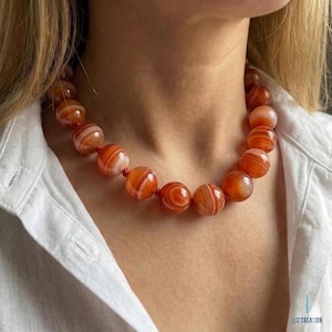 May include: A necklace featuring large, round beads in shades of orange and white. The beads are strung together on a red cord, creating a bold and eye-catching accessory. The necklace is worn against a white shirt.