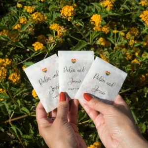 May include: Three white paper bags with rainbow hearts and the text "Robin and Josie" written in black calligraphy. The bags are held in the hands of a person.