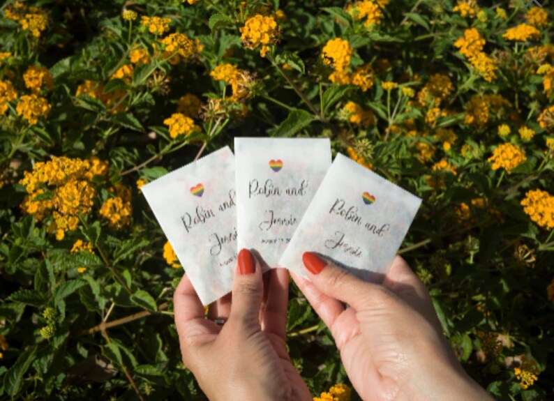 May include: Three white paper bags with rainbow hearts and the text "Robin and Josie" written in black calligraphy. The bags are held in the hands of a person.