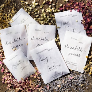 May include: White paper bags with black text, each with a different name and date. The bags are filled with flower petals and are scattered on a wooden surface. The text on the bags reads: "Toss me... XOXO Elizabeth + James", "Sprinkle some... XOXO Amy & Andrew", "Toss me... XOXO Elizabeth + James", "Toss me... XOXO Elizabeth + James", "Sprinkle some... XOXO Amy & Andrew".