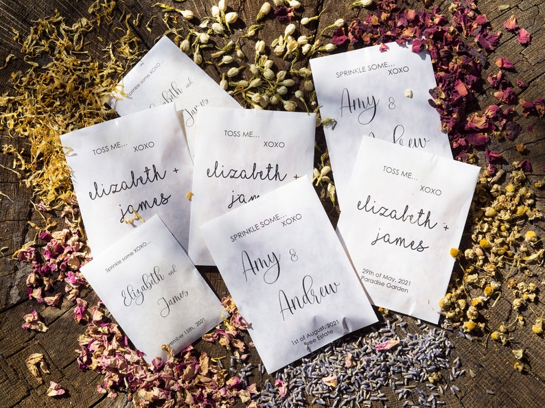 May include: White paper bags with black text, each with a different name and date. The bags are filled with flower petals and are scattered on a wooden surface. The text on the bags reads: "Toss me... XOXO Elizabeth + James", "Sprinkle some... XOXO Amy & Andrew", "Toss me... XOXO Elizabeth + James", "Toss me... XOXO Elizabeth + James", "Sprinkle some... XOXO Amy & Andrew".