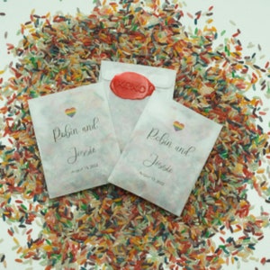May include: White paper bags with a rainbow heart and the text "Robin and Jessie" with the date August 13, 2023. The bags are filled with colorful rice.