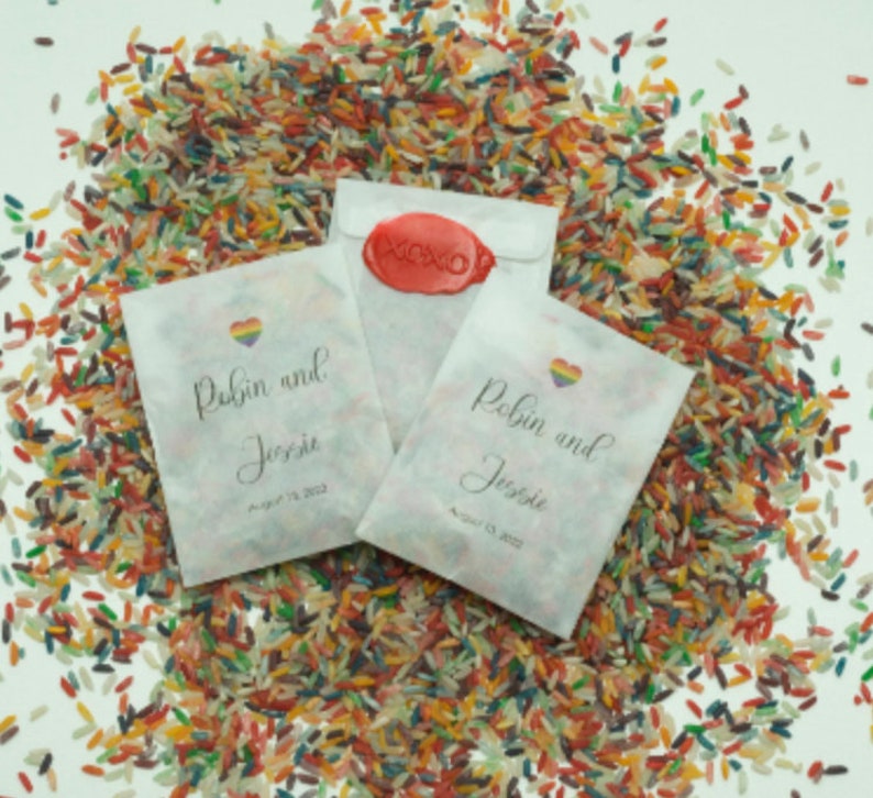 May include: White paper bags with a rainbow heart and the text "Robin and Jessie" with the date August 13, 2023. The bags are filled with colorful rice.