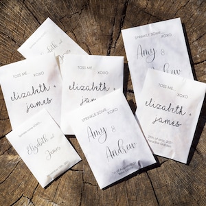 May include: Six white paper envelopes with black text. The envelopes are for wedding favors and have the names of the couples and the date of their wedding. The text on the envelopes reads: "Toss Me... XOXO Elizabeth + James", "Sprinkle Some... XOXO Elizabeth + James", "Toss Me... XOXO Elizabeth + James", "Sprinkle Some... XOXO Amy & Andrew", "Sprinkle Some... XOXO Amy & Andrew", and "Toss Me... XOXO Elizabeth + James".
